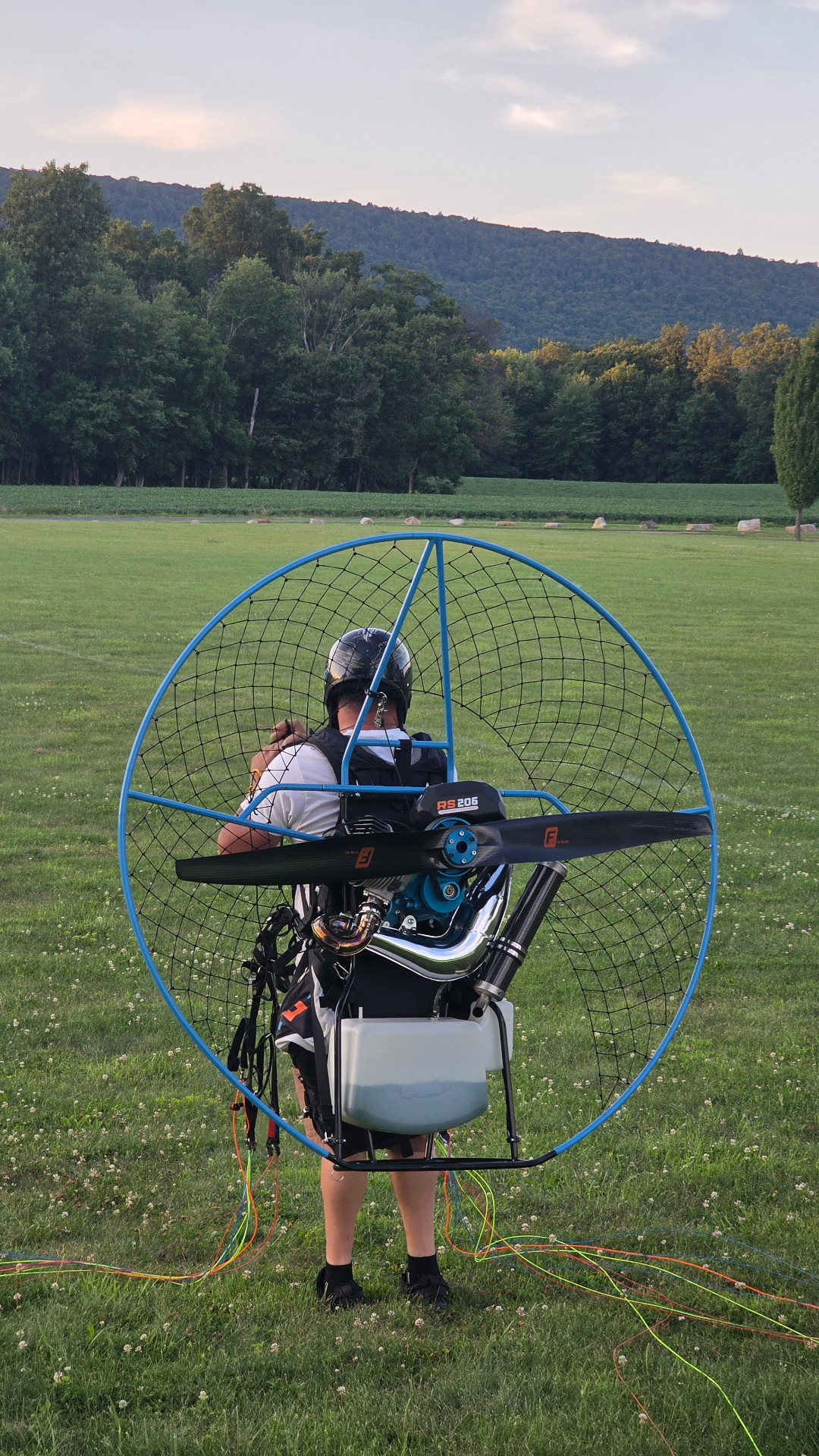 Pilot geared up with a blue-hoop HE RS 206 paramotor in a mountain field